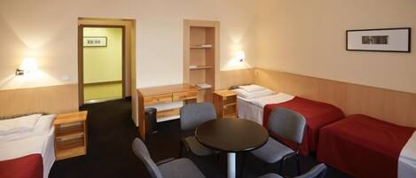A hotel room with two single beds, a small round table, and two chairs. There is a desk with a chair and a shelf unit.