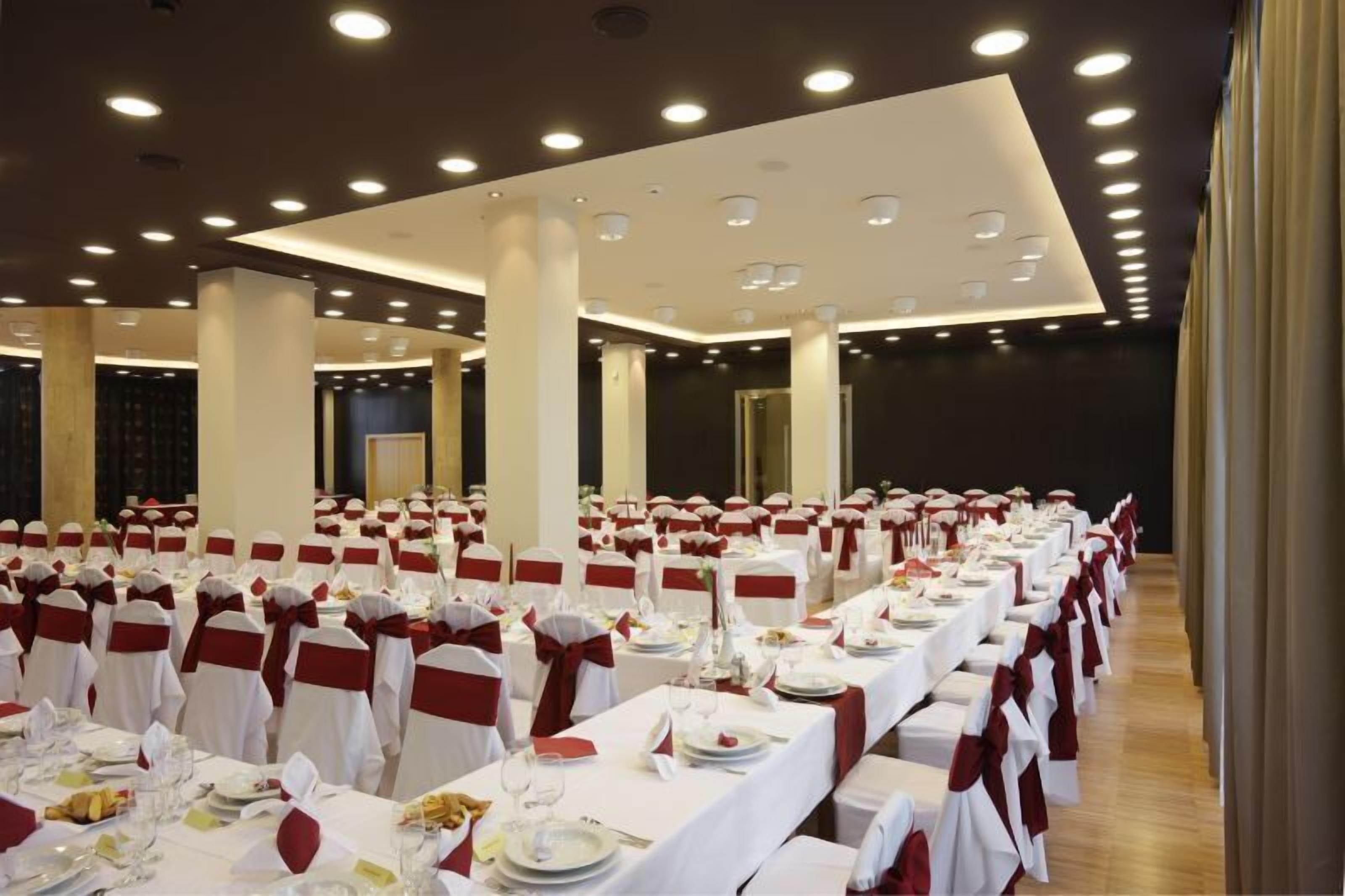 Una sala da banchetto con file di tavoli apparecchiati per un evento formale, con tovaglie bianche e nastri rossi.