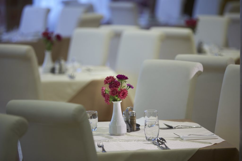 Une table à manger dressée avec un vase blanc rempli de fleurs, des verres à eau et des couverts.