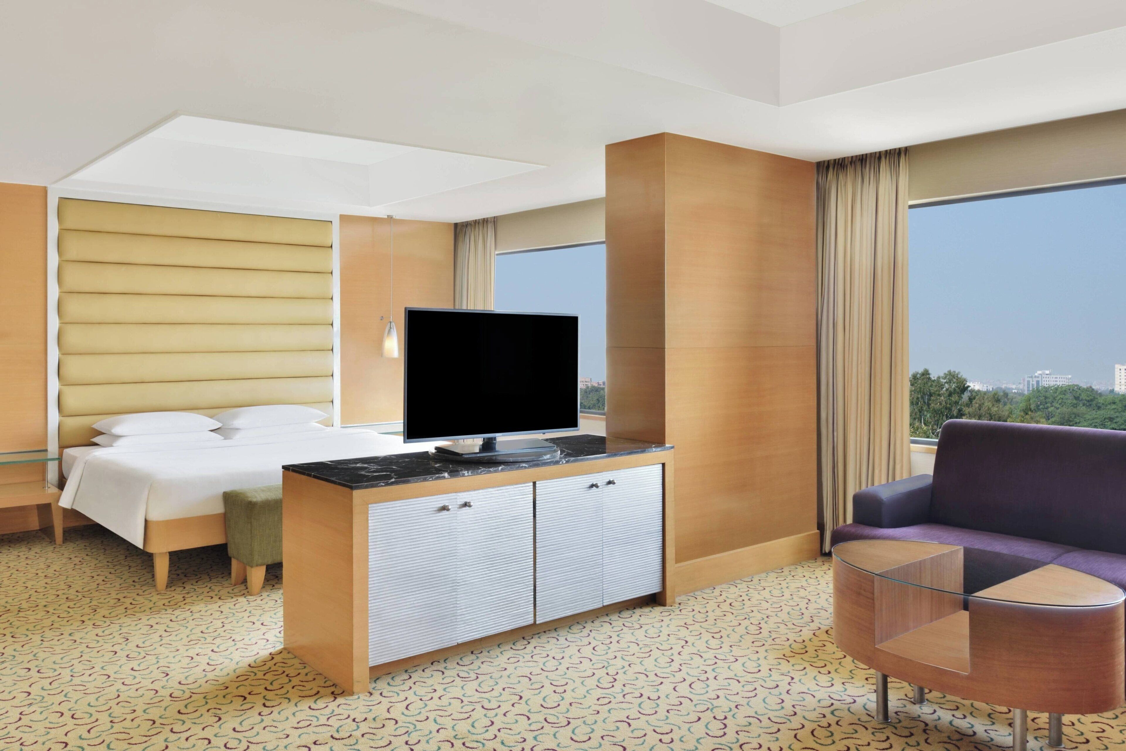 Habitación de hotel con cama, televisor sobre un mueble de madera, sofá y una pequeña mesa de vidrio.
