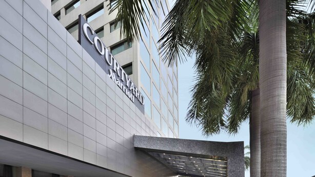 Entrada de um hotel Courtyard Marriott com galeria coberta, palmeiras e área ajardinada.
