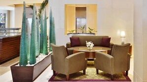 Um saguão com balcão de recepção, área de estar com sofá e cadeiras, e pilares de vidro decorativos.