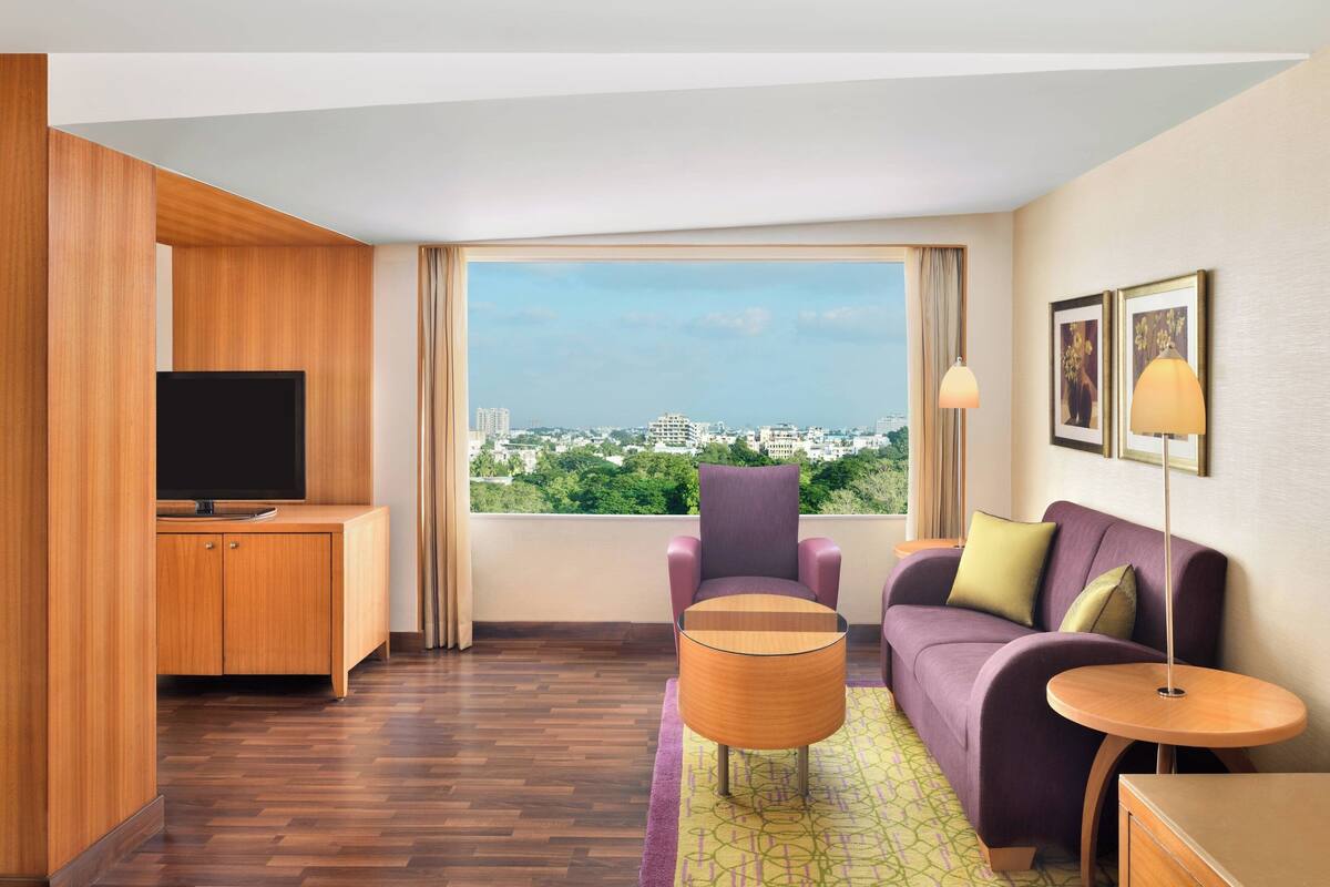 Une chambre d’hôtel moderne dotée d’une grande fenêtre, d’un téléviseur à écran plat, d’un canapé violet, d’une armoire en bois et d’une petite table ronde.