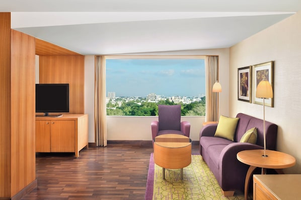 Una habitación de hotel moderna con un ventanal grande, un televisor de pantalla plana, un sofá morado, un armario de madera y una mesita redonda.