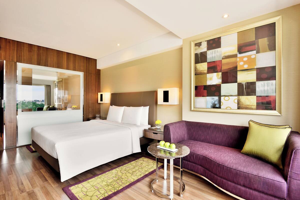 Une chambre d’hôtel avec un lit, un canapé, une petite table avec des pommes, un miroir et un grand tableau abstrait accroché au mur.
