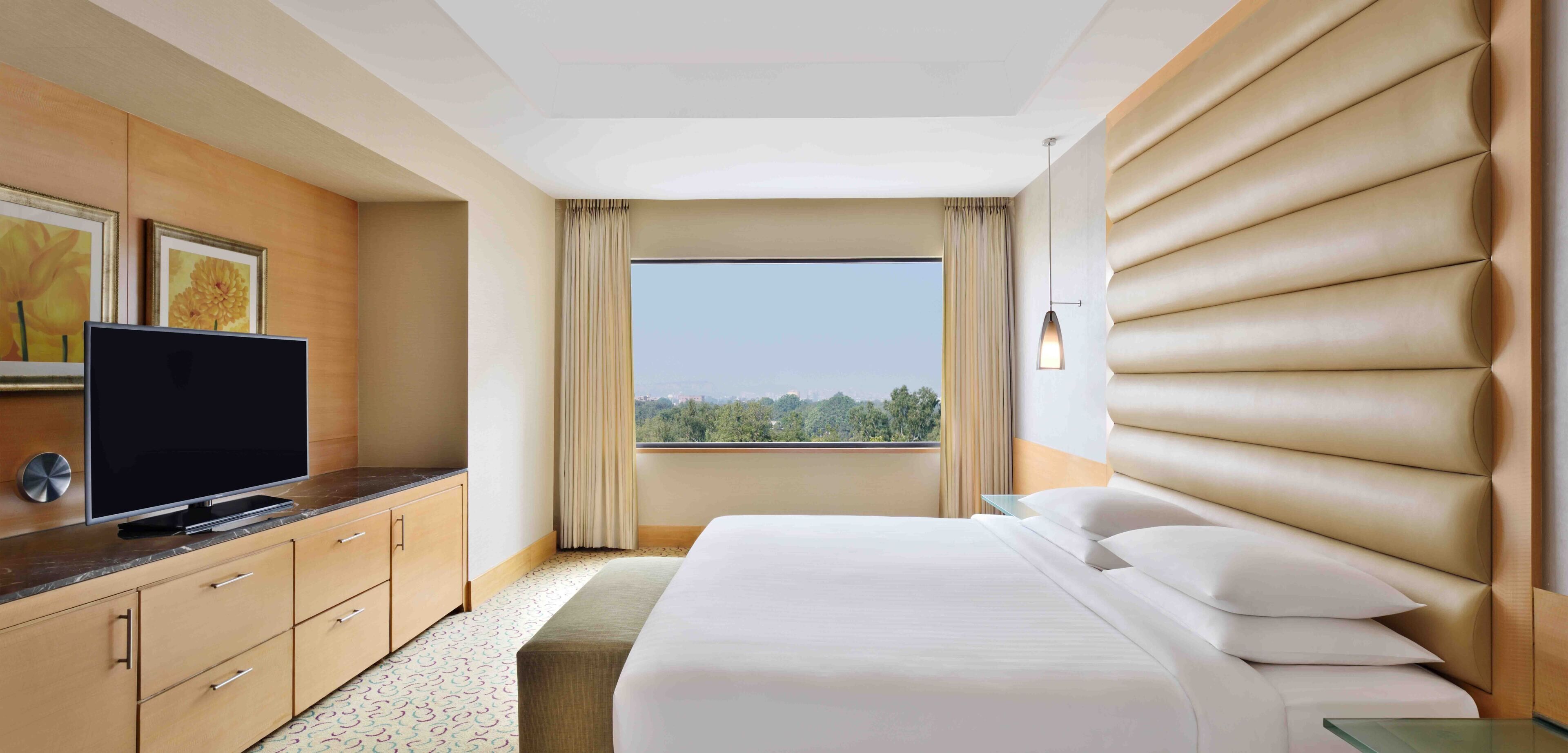 A hotel room with a bed, a TV on a wooden dresser, a large window with a view of trees, and a ceiling with a unique design.
