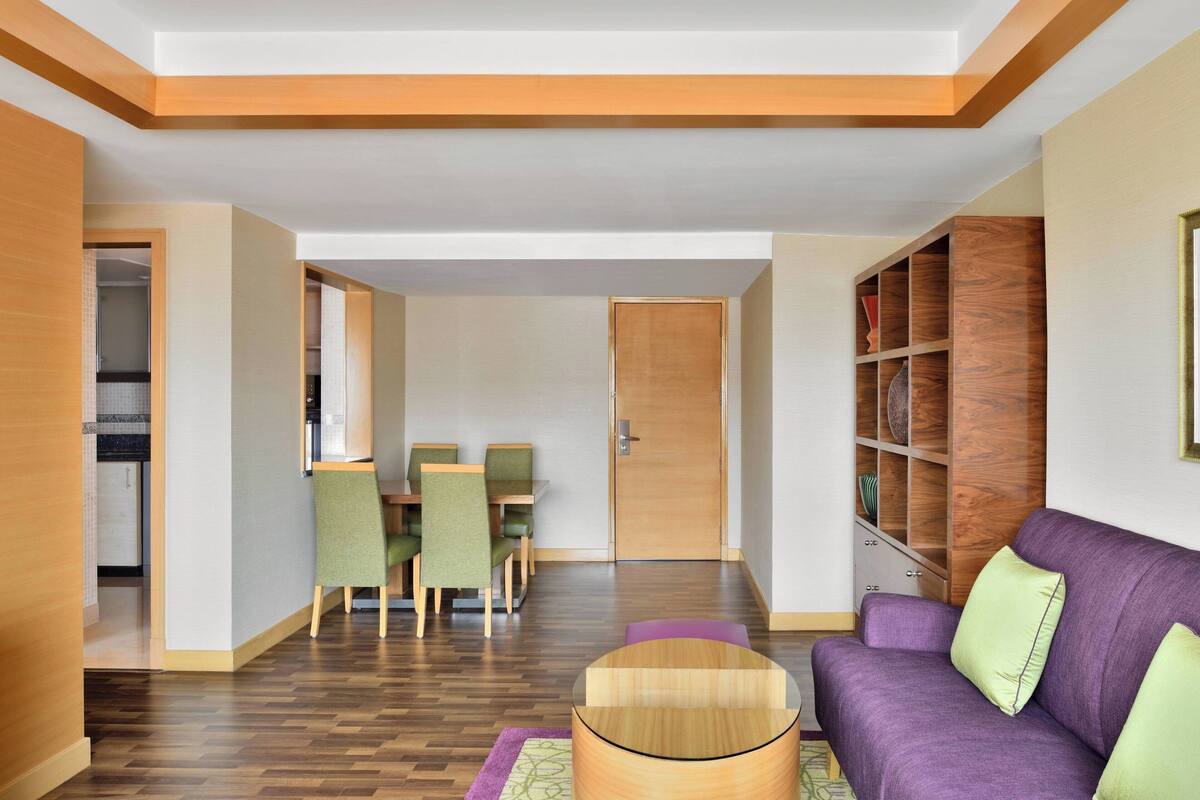 Un salon moderne avec un canapé violet, des fauteuils verts, une table basse en bois et une bibliothèque en bois.