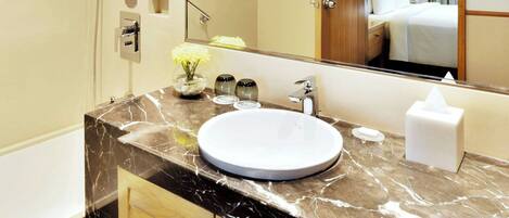 A modern bathroom with a marble vanity, a large mirror, a shower, and a visible bedroom in the reflection.