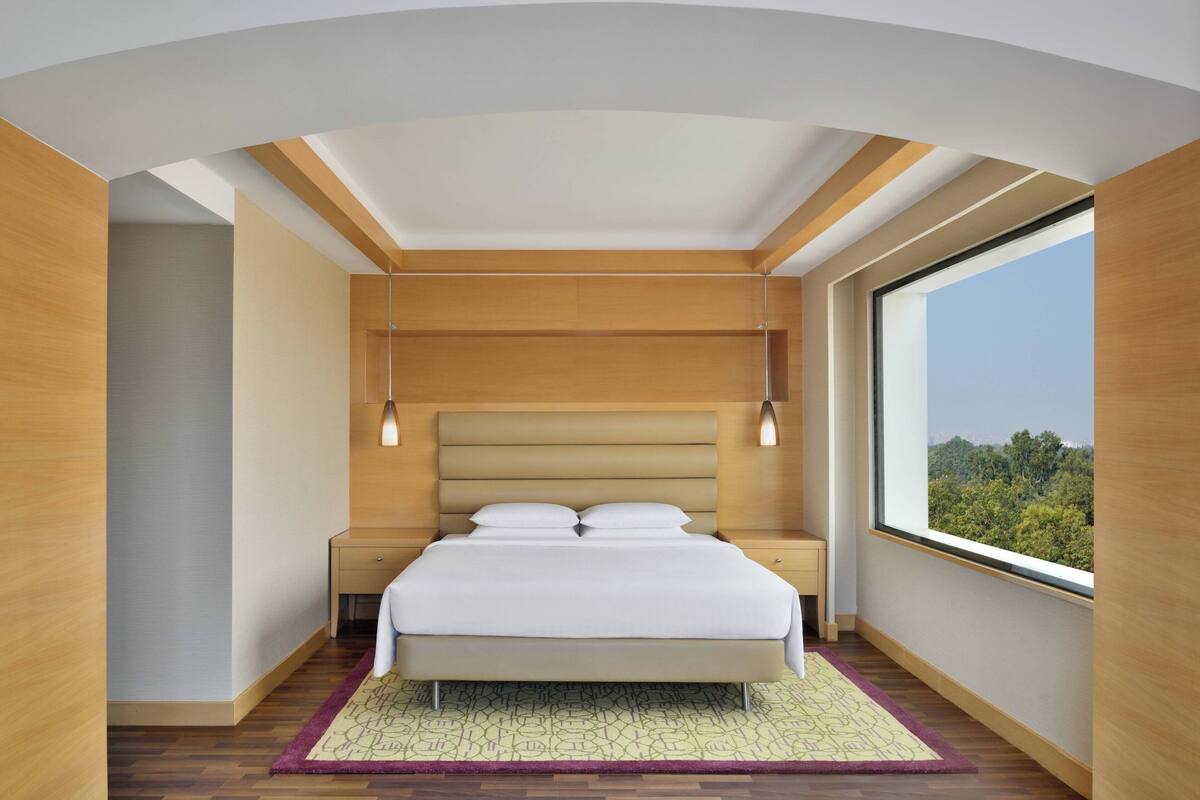 Une chambre moderne avec un grand lit, une tête de lit en bois, des tables de chevet et une grande fenêtre donnant sur un paysage verdoyant.