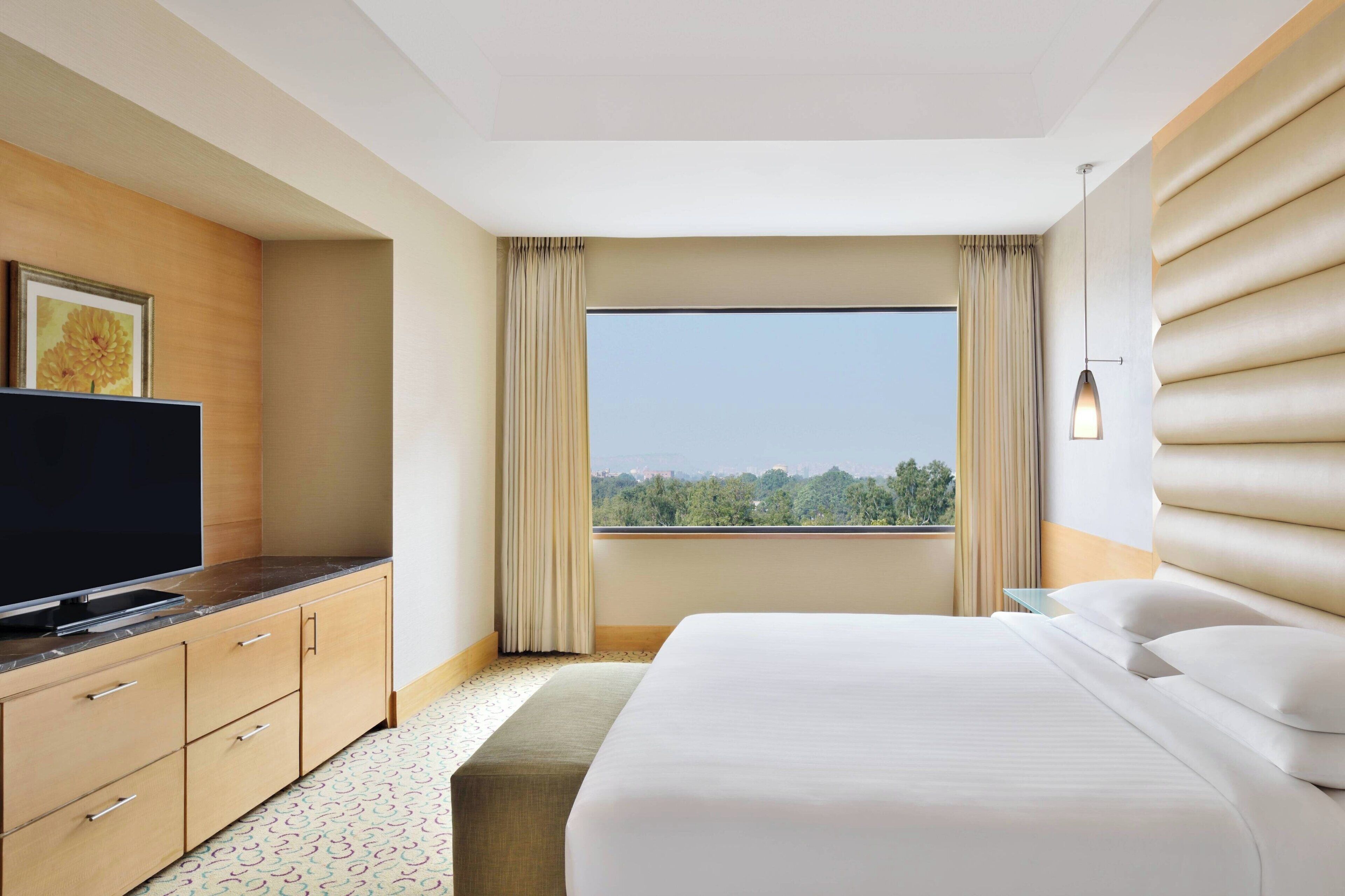 A hotel room with a bed, a TV on a wooden cabinet, a window with curtains, and a view of greenery.
