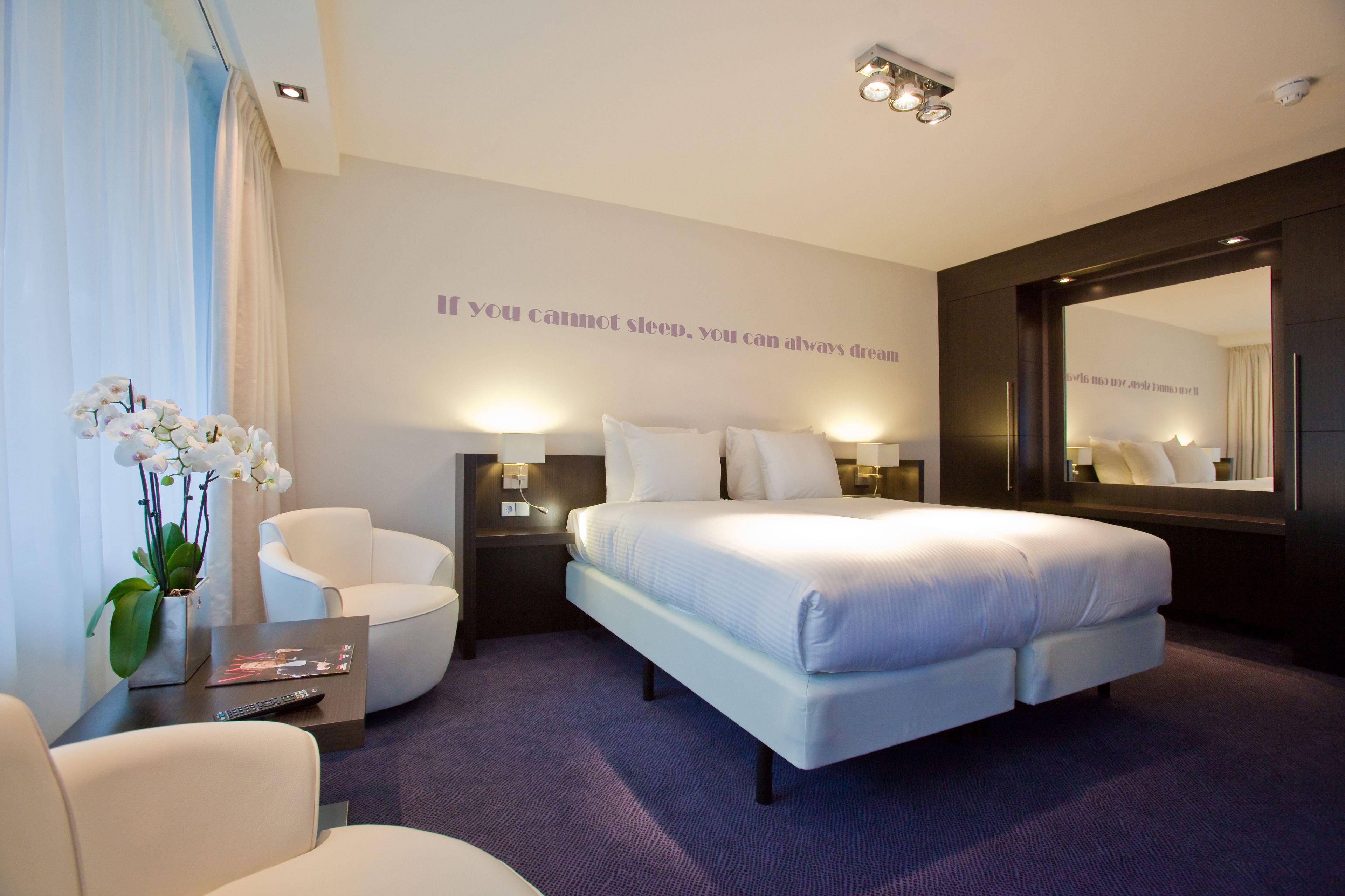 Una habitación de hotel moderna con una cama grande, una zona de estar con una silla, una mesita de noche y un espejo.