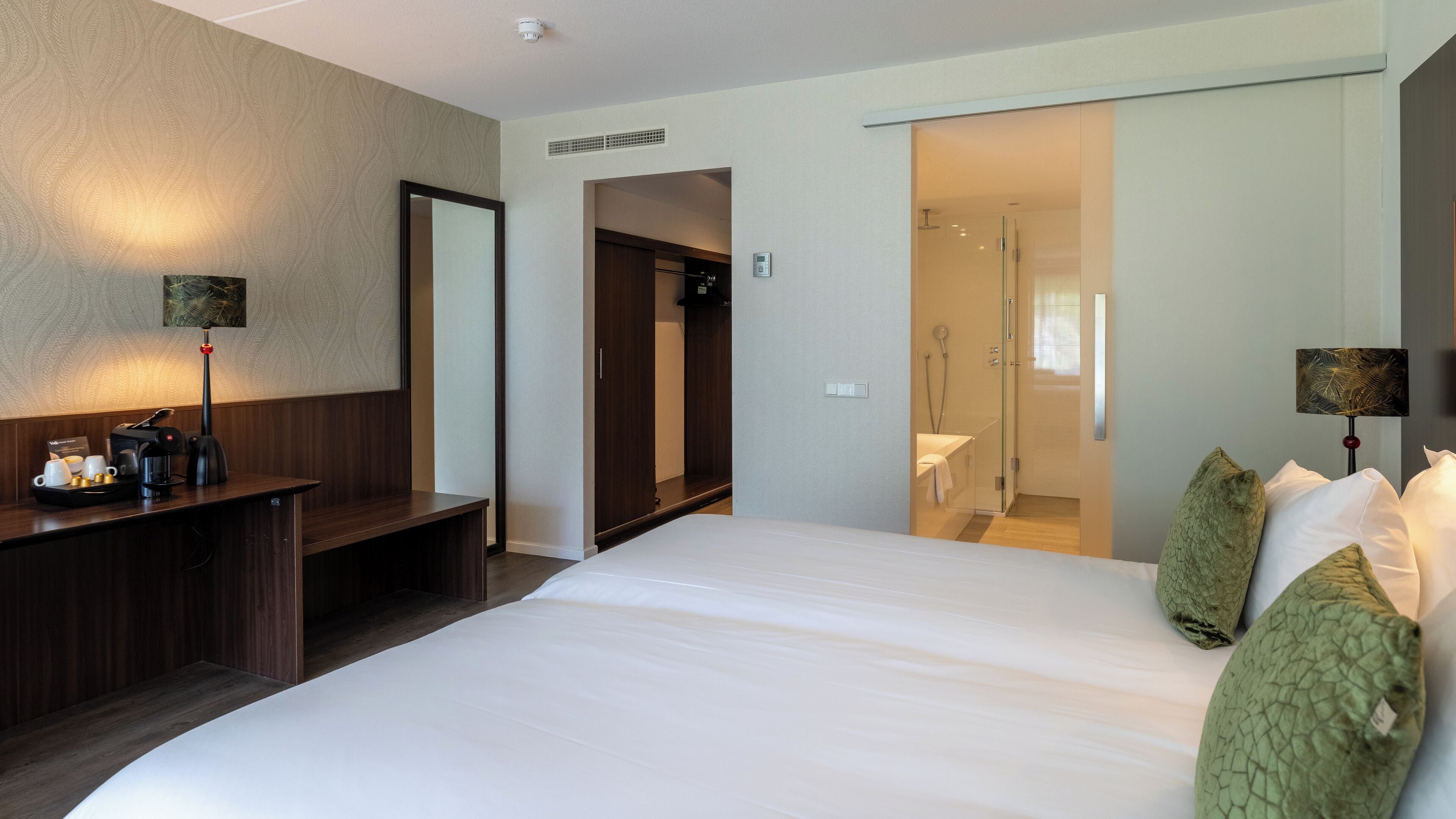 A hotel room with a large bed, a desk with a coffee maker, a mirror, and a shower area.