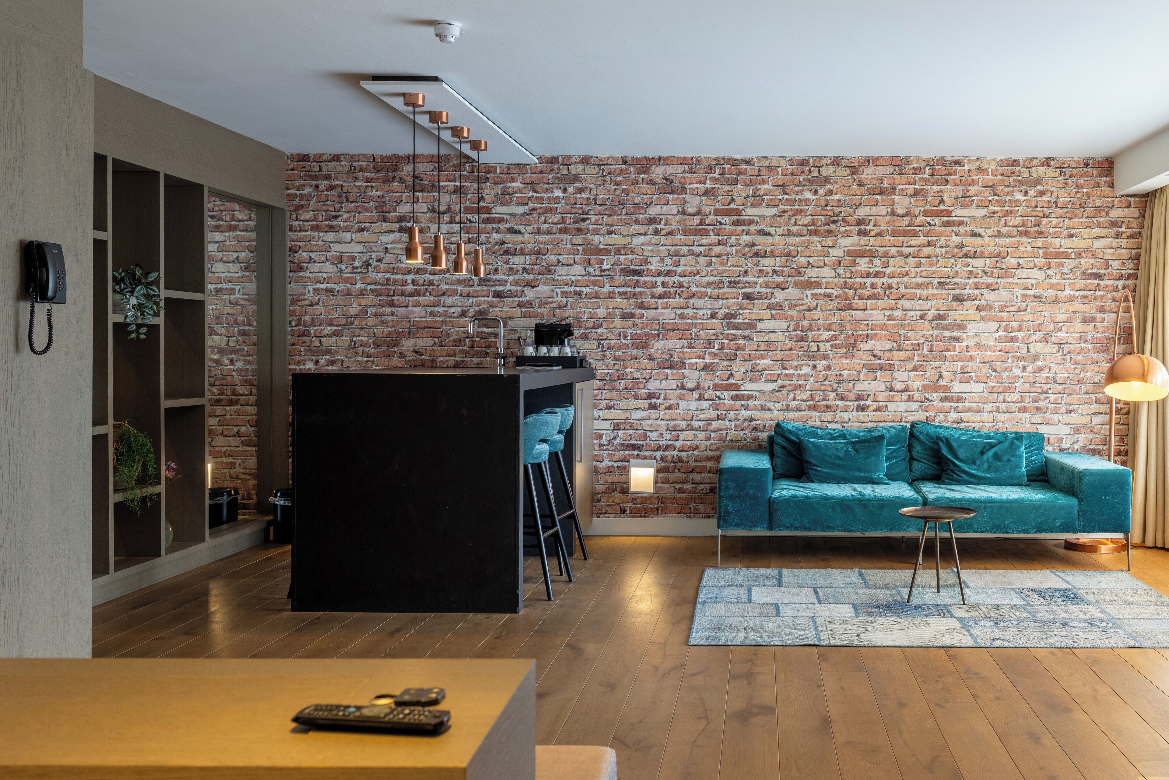 A modern living space with a brick wall, a black kitchen island, a teal sofa, and a wooden floor.