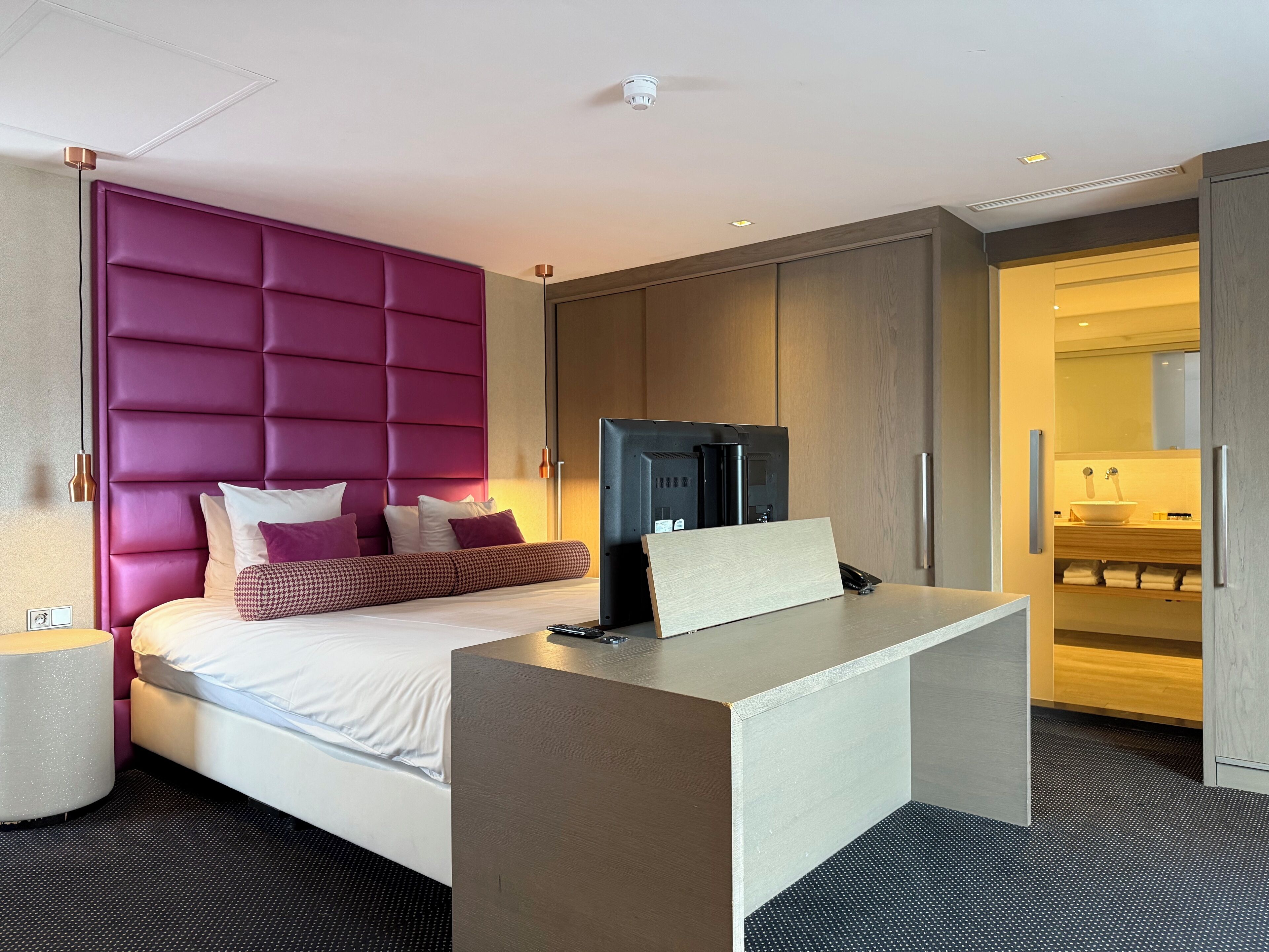 A modern hotel room with a large bed, a desk, and a bathroom visible through an open door.