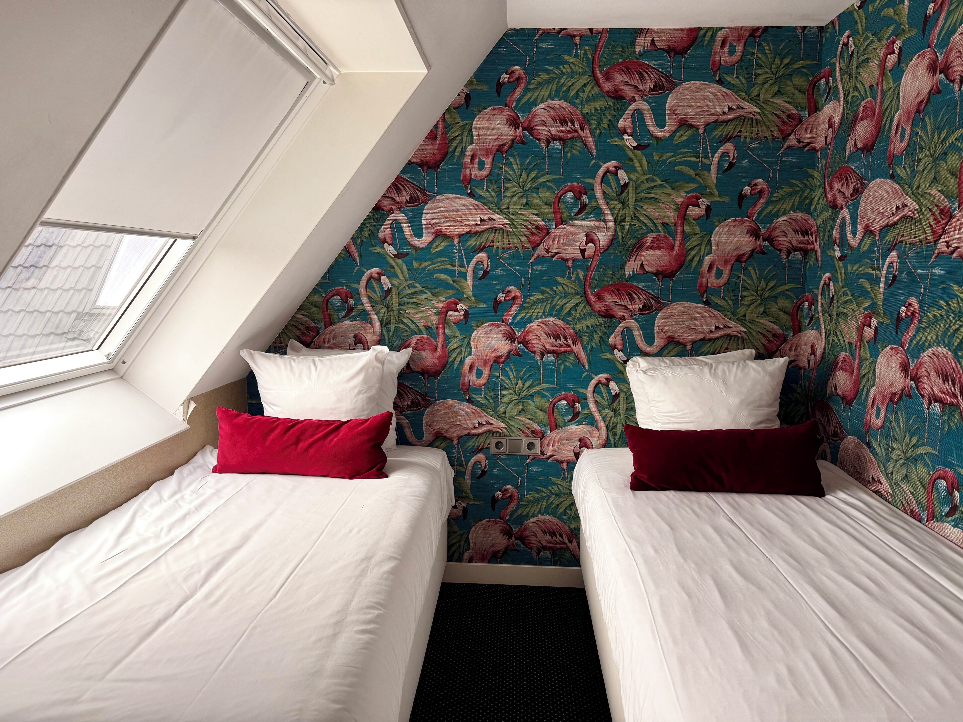 A room with two beds, each with white bedding and red pillows, against a wall with a flamingo pattern. A skylight is visible above the beds.
