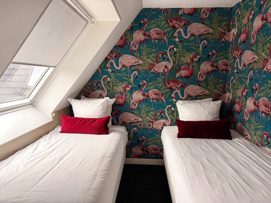 A room with two beds, each with white bedding and red pillows, against a wall with a flamingo pattern. A skylight is visible above the beds.