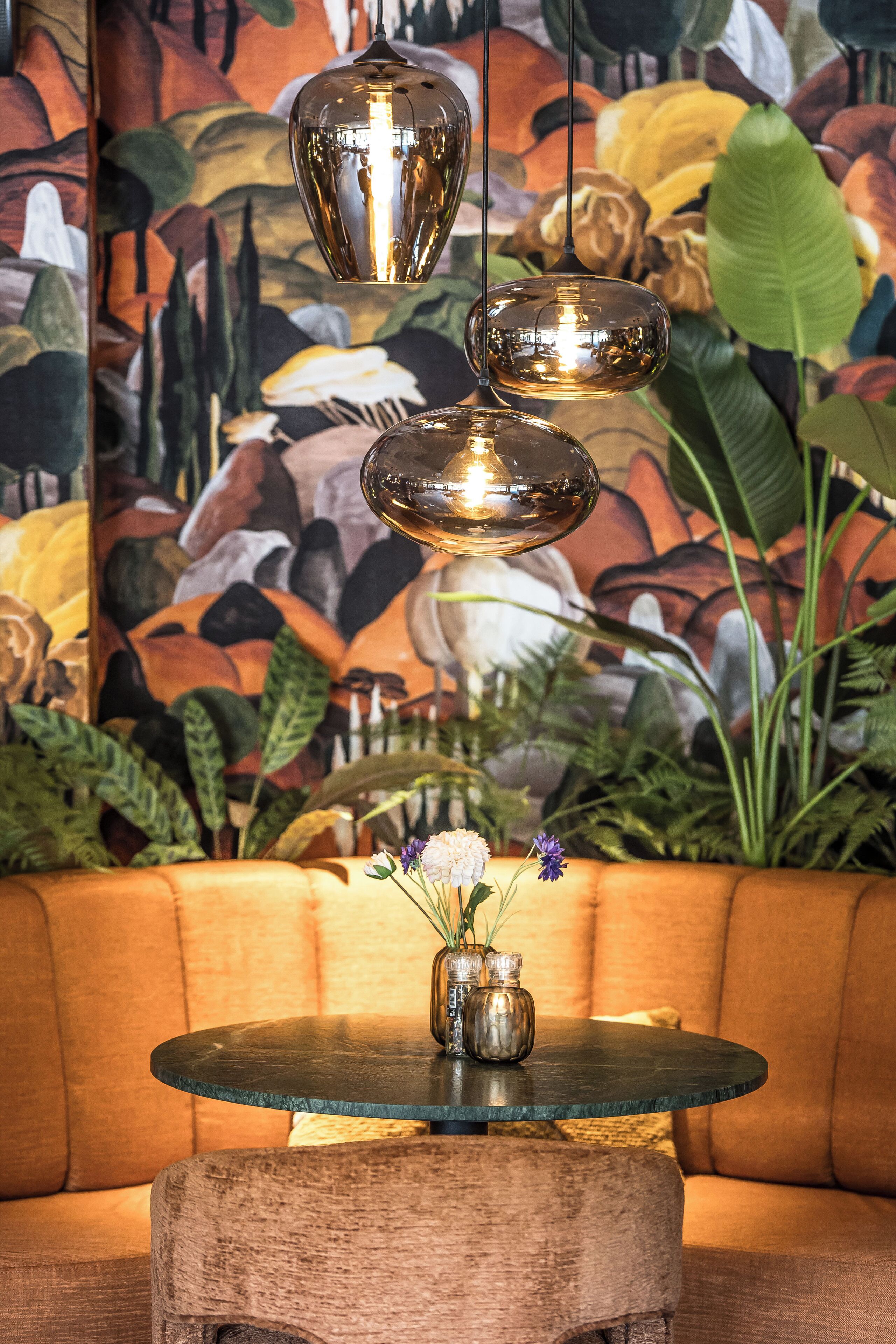 A round glass table with a metal vase and flowers, a wooden stool, and a sofa in front of a mural with plants and flowers.