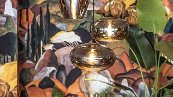 A round glass table with a metal vase and flowers, a wooden stool, and a sofa in front of a mural with plants and flowers.