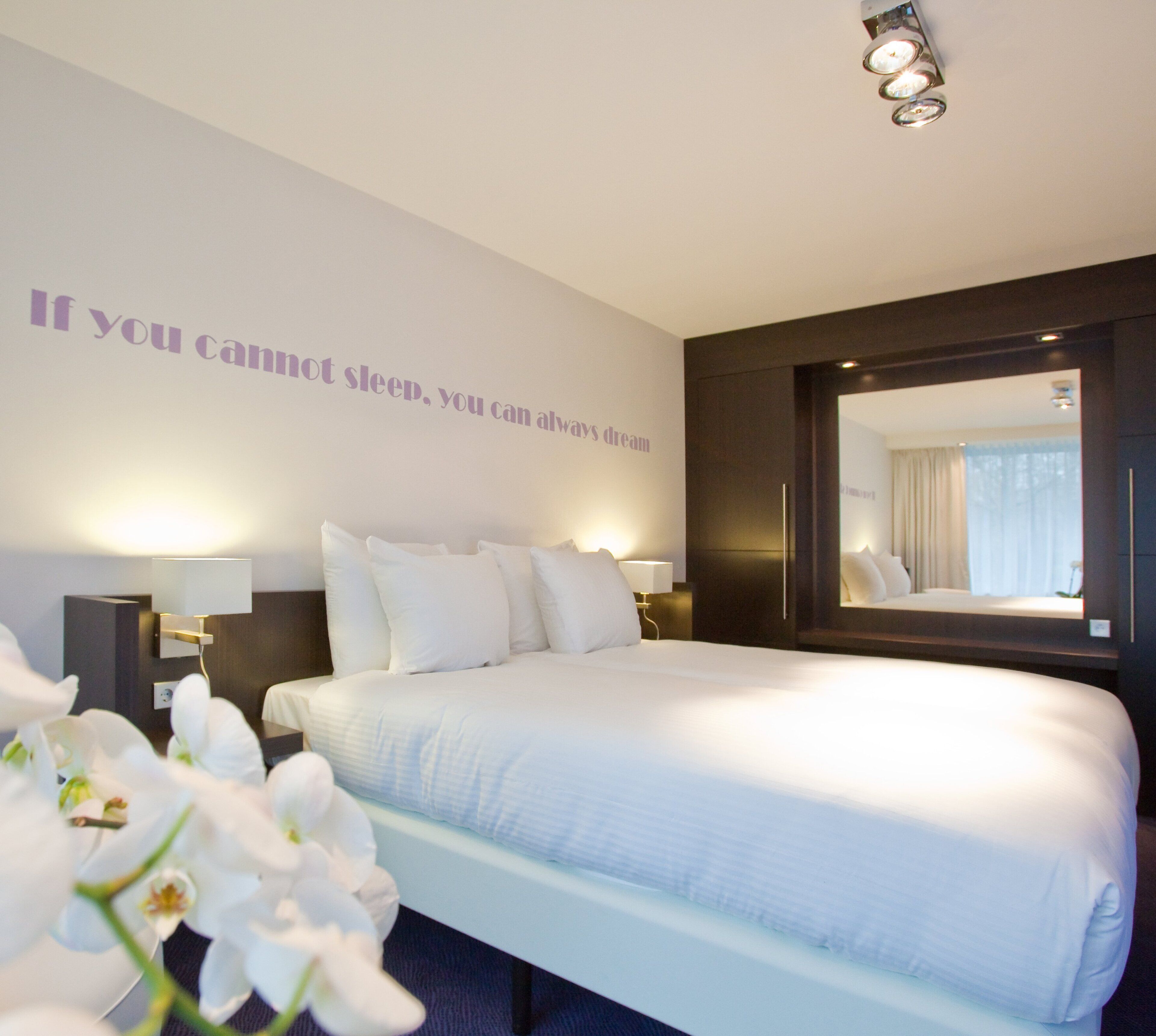 A hotel room with a large bed, white bedding, a wall-mounted mirror, and a motivational quote.