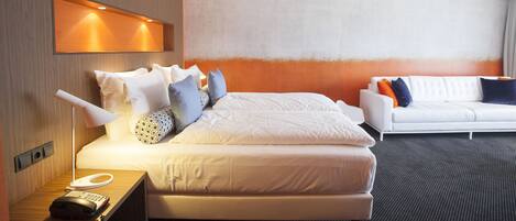 Quarto de hotel com cama, mesa de cabeceira, luminária e sofá branco.