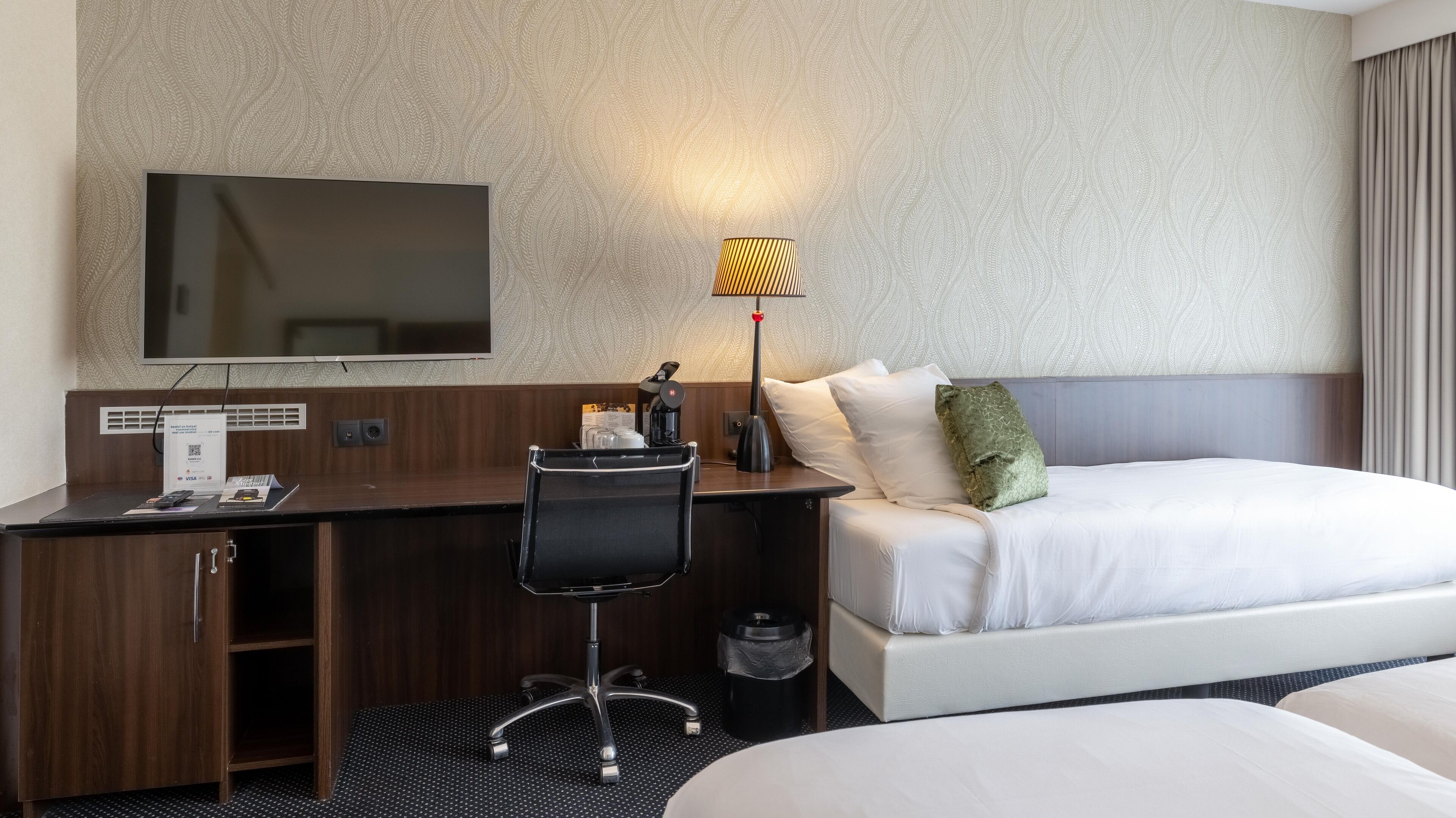 Ein Hotelzimmer mit einem Bett, einem Schreibtisch mit Stuhl, einem an der Wand befestigten Fernseher und einer Lampe.