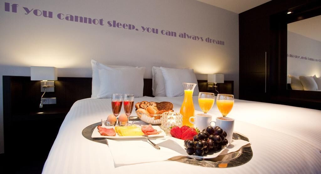 A neatly made bed with a breakfast tray, including eggs, toast, fruit, and juice, in a hotel room with a motivational quote on the wall.