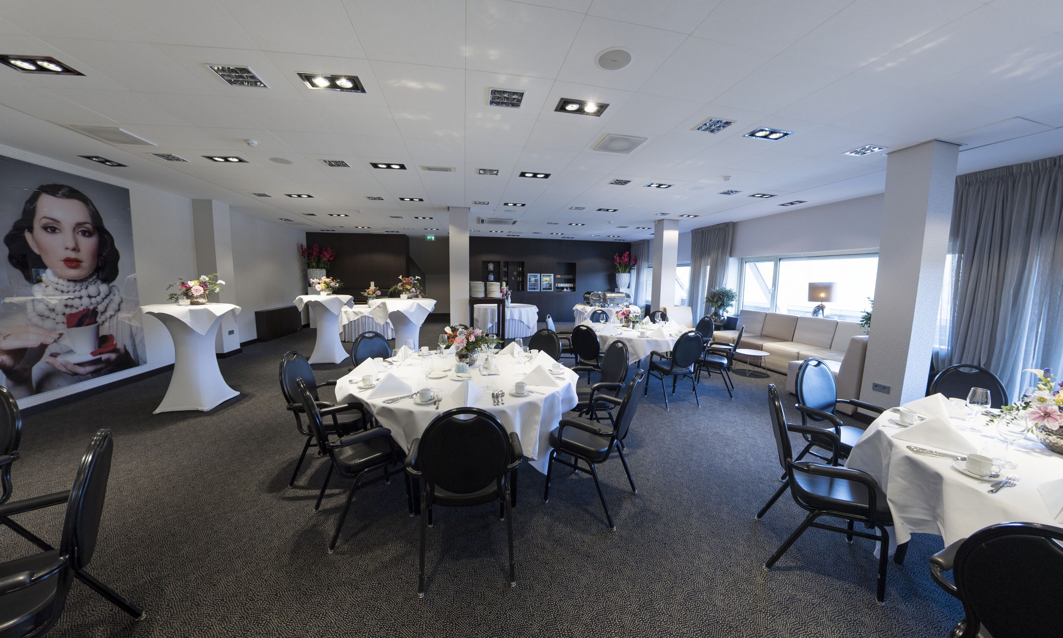 A hotel dining area with round tables, black chairs, and white tablecloths.