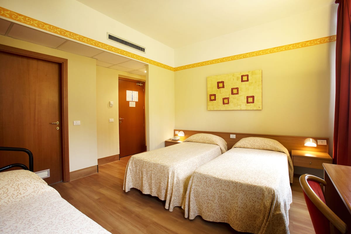 A hotel room with two single beds, a wooden door, a small bedside table with a lamp, and a wall-mounted artwork.