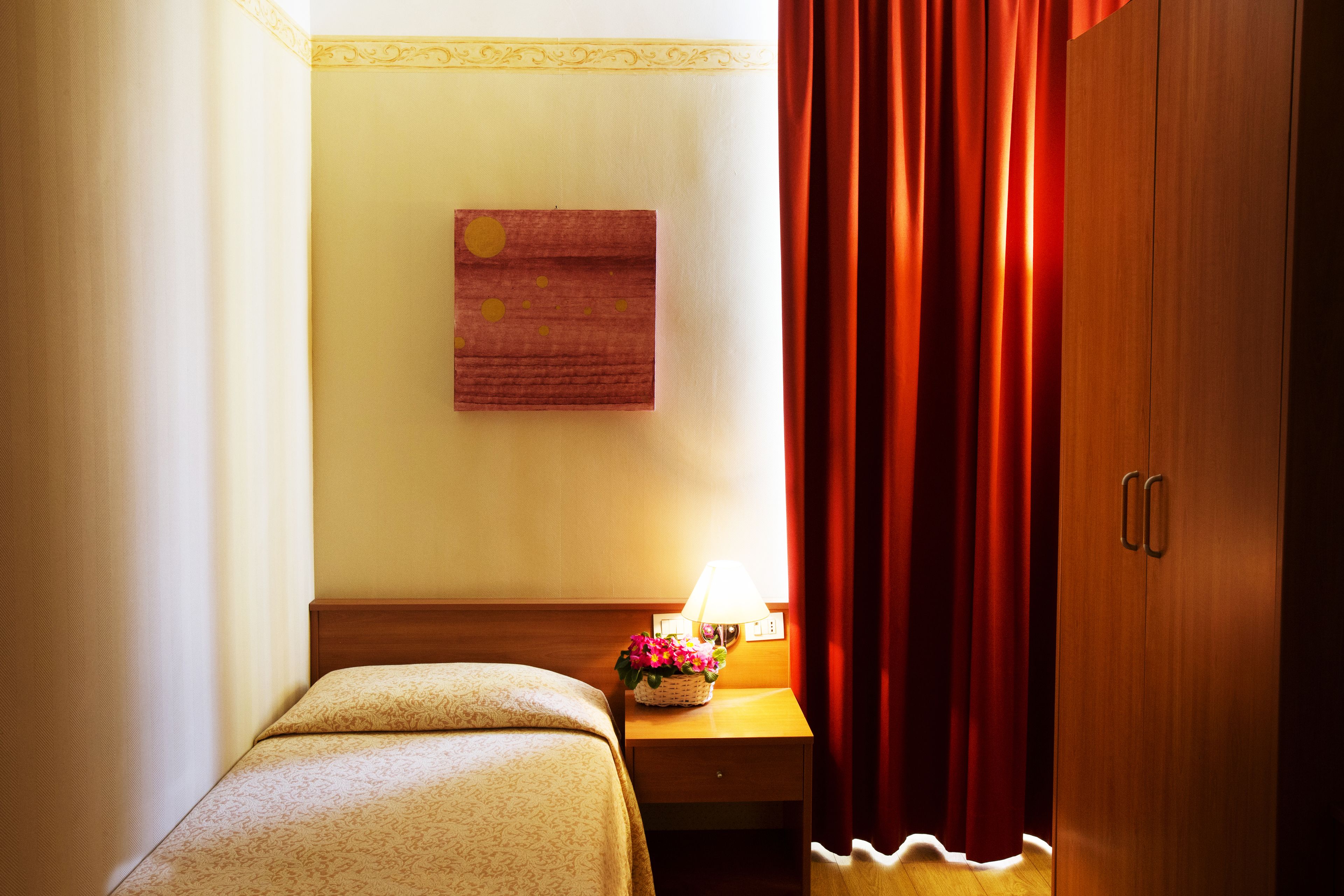 Habitación de hotel con cama, mesita de noche con lámpara, armario de madera y una obra de arte colgada en la pared.