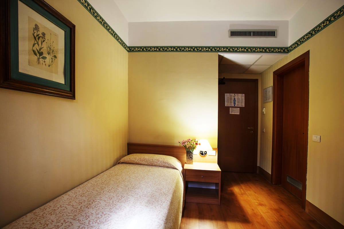 A hotel room with a bed, a nightstand with a lamp, a framed botanical print on the wall, and a wooden door.