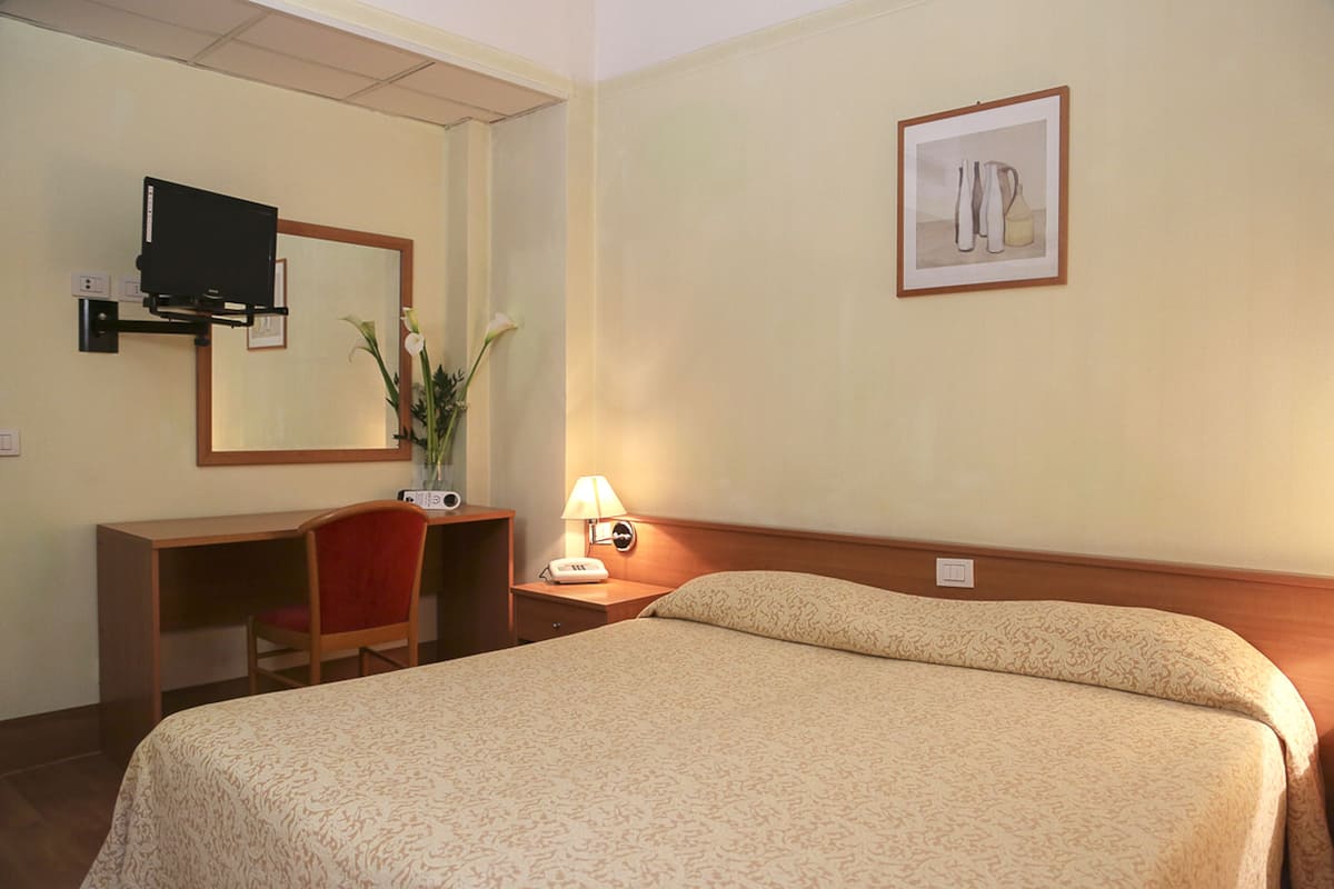 A hotel room with a bed, a desk, a chair, a lamp, a mirror, and a framed picture on the wall.
