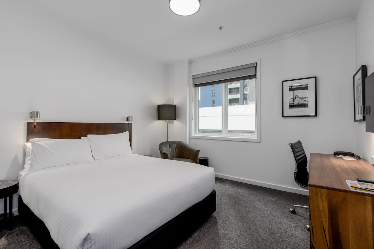 A hotel room with a large bed, a desk, a chair, a lamp, and a window with a view of buildings.