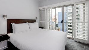 Habitación de hotel con una cama grande, cabecera de madera, mesita de noche, lámpara de pared y puerta corrediza de vidrio que da a un balcón con vistas a la ciudad.