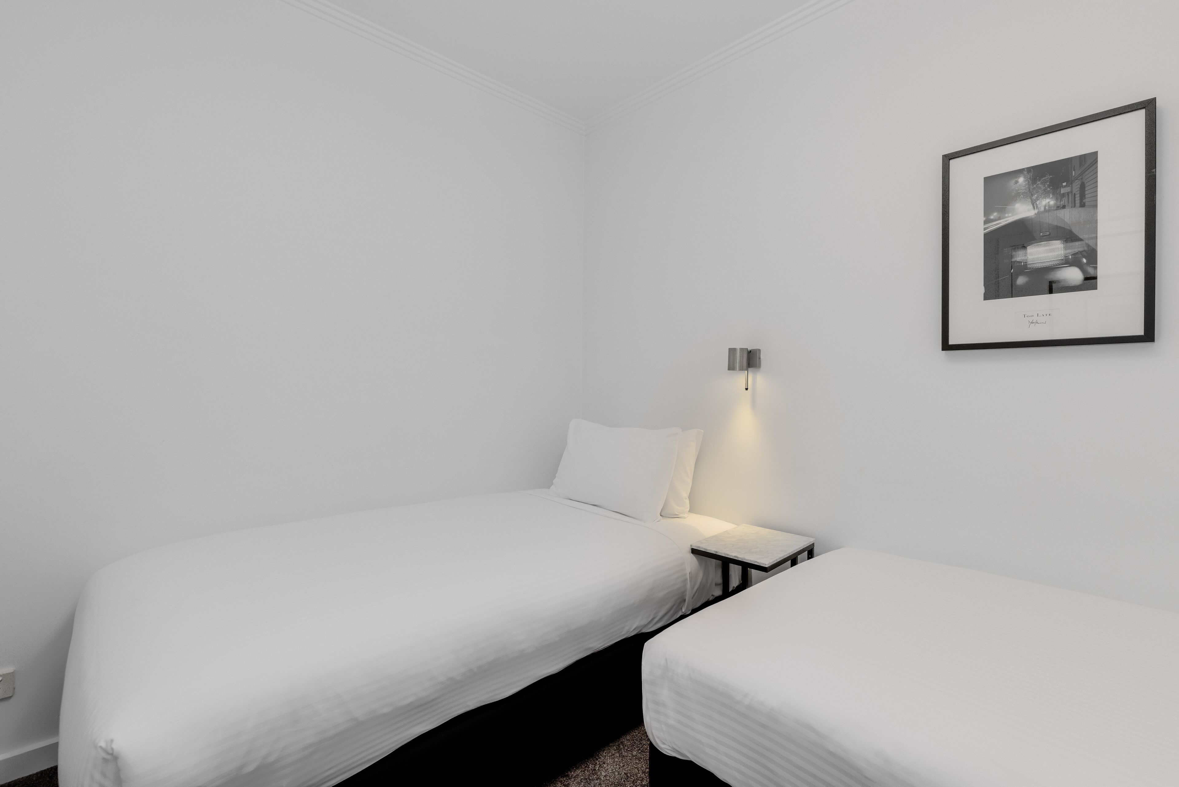 Ein schlicht eingerichtetes Zimmer mit zwei Einzelbetten, einem Nachttisch mit Lampe und einem gerahmten Foto an der Wand.