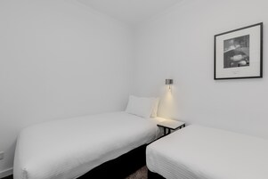 Una habitación minimalista con dos camas individuales, una mesita de noche con lámpara, y una fotografía enmarcada en la pared.