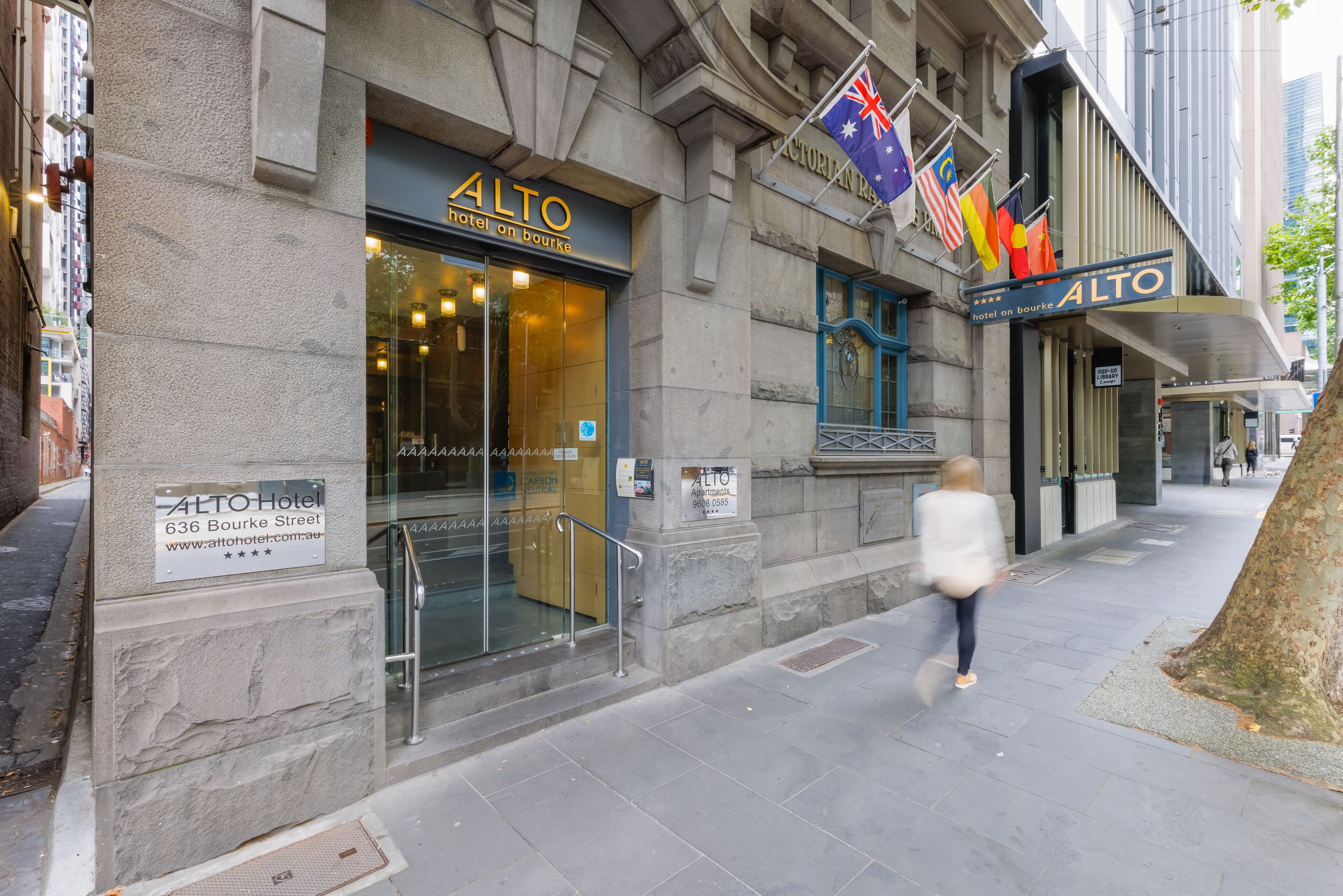 The exterior of the Alto Hotel with a stone facade, large glass entrance, and multiple flags.