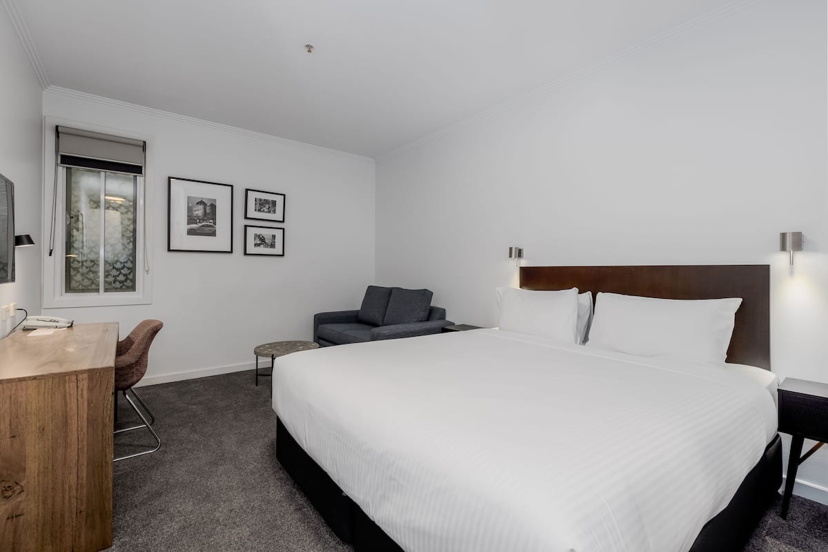 A hotel room with a large bed, a desk, a chair, and a sofa. There are framed pictures on the wall and a window with a view.
