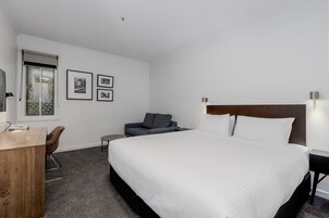 A hotel room with a large bed, a desk, a chair, and a sofa. There are framed pictures on the wall and a window with a view.