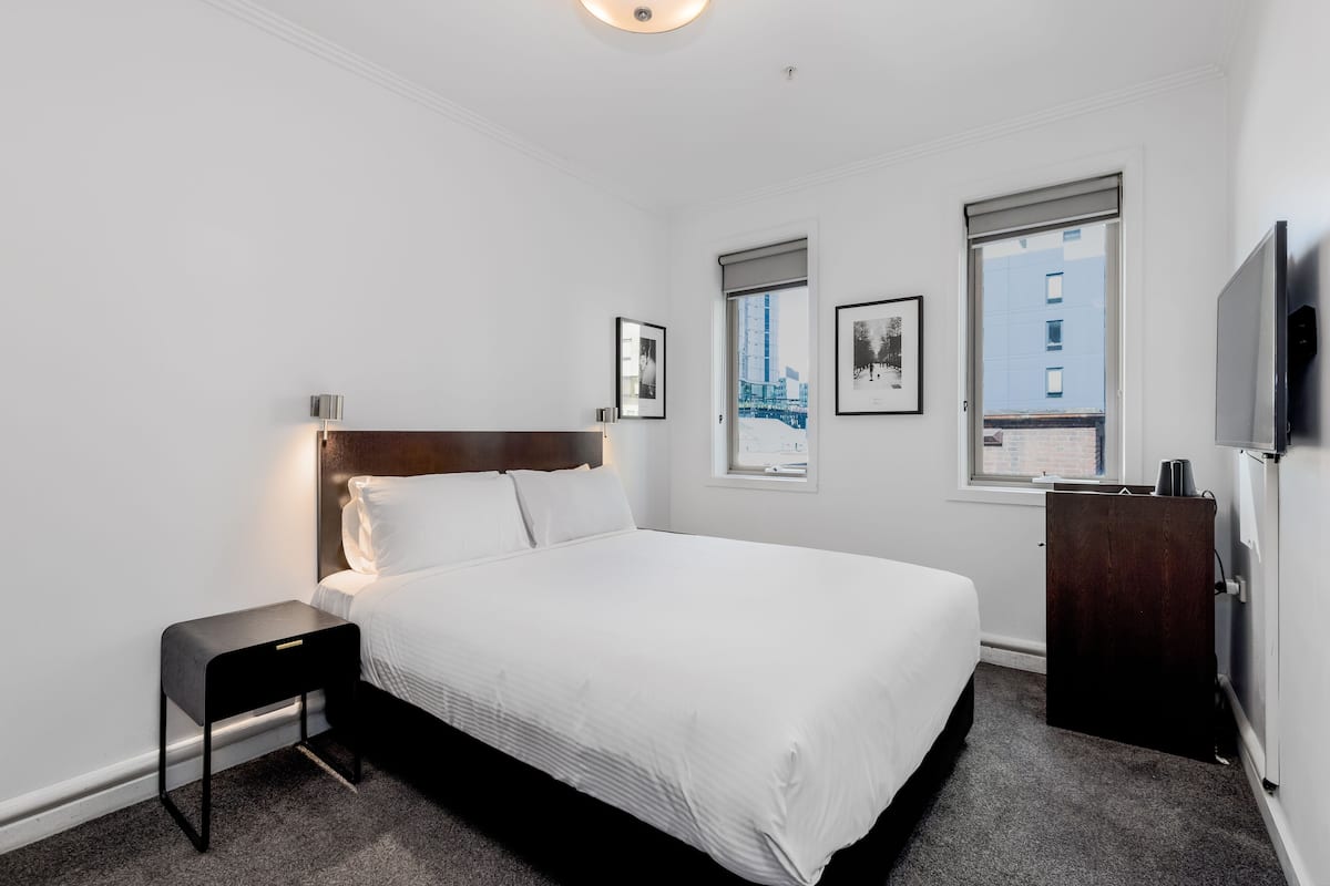 A hotel room with a large bed, a bedside table, a TV, and a window with a view of a building.