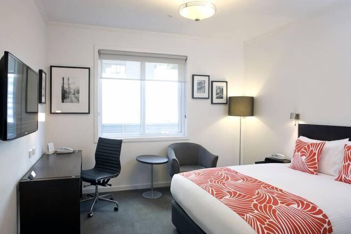 A hotel room with a bed, desk, chair, small table, lamp, and framed pictures on the wall.