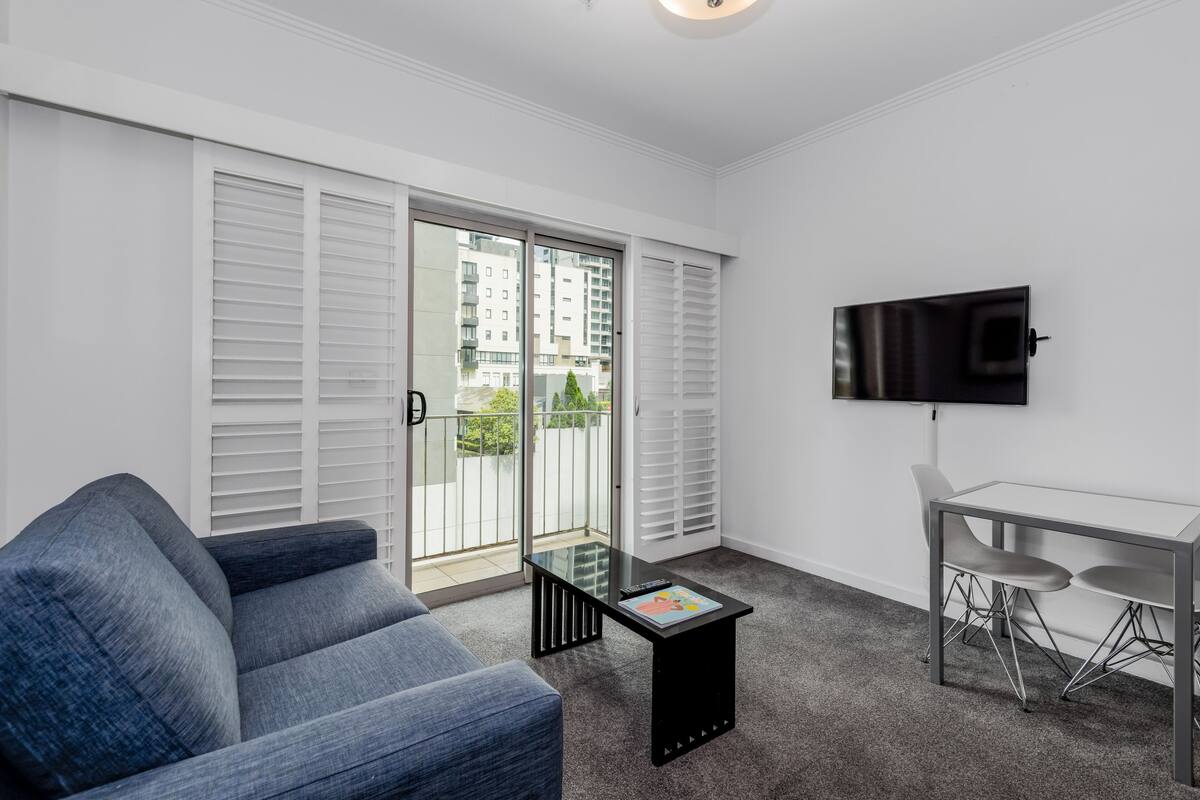 A modern living room with a sofa, a coffee table, a TV mounted on the wall, and a balcony with white shutters.
