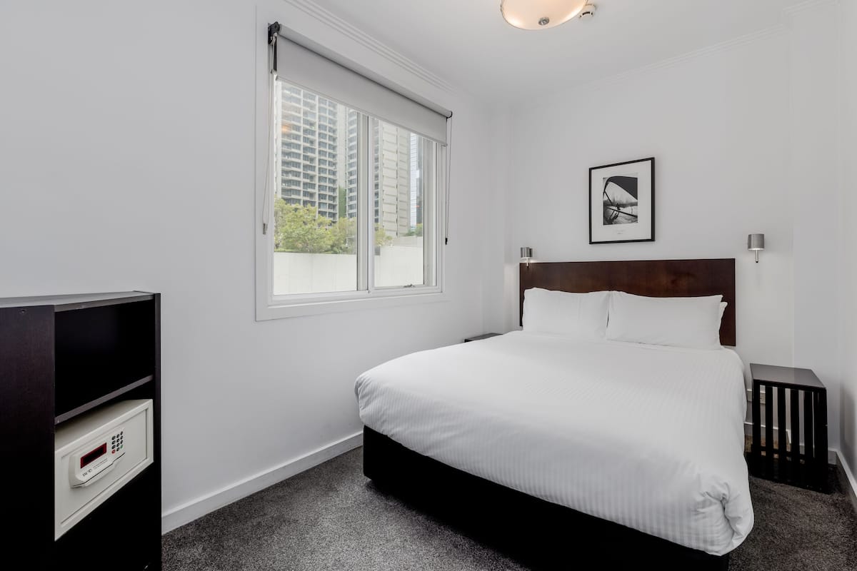 A hotel room with a large bed, a window with blinds, a bedside table, a wall-mounted picture, and a built-in safe.