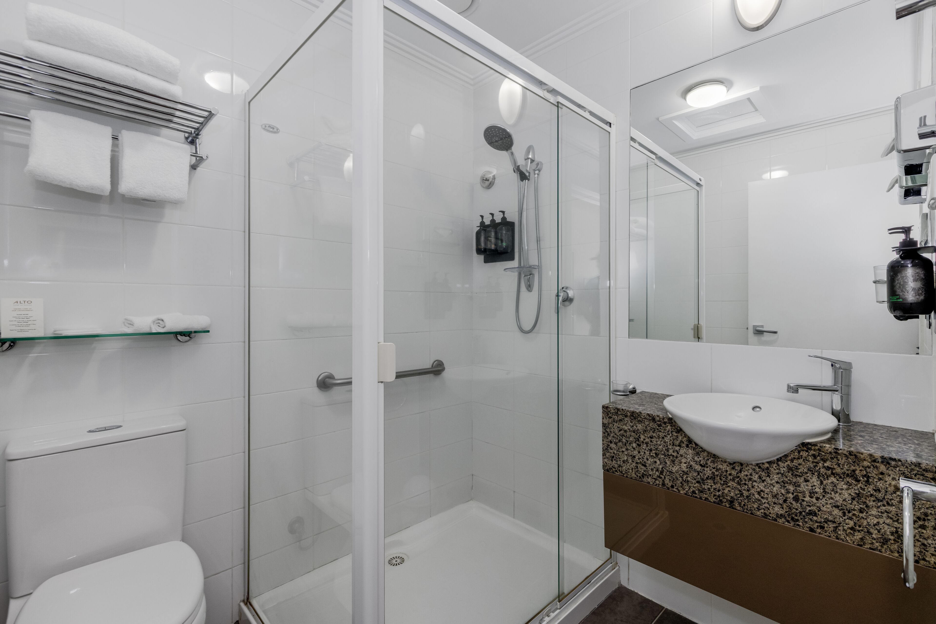 A modern bathroom with a glass-enclosed shower, a white toilet, a granite countertop sink, and a large mirror.