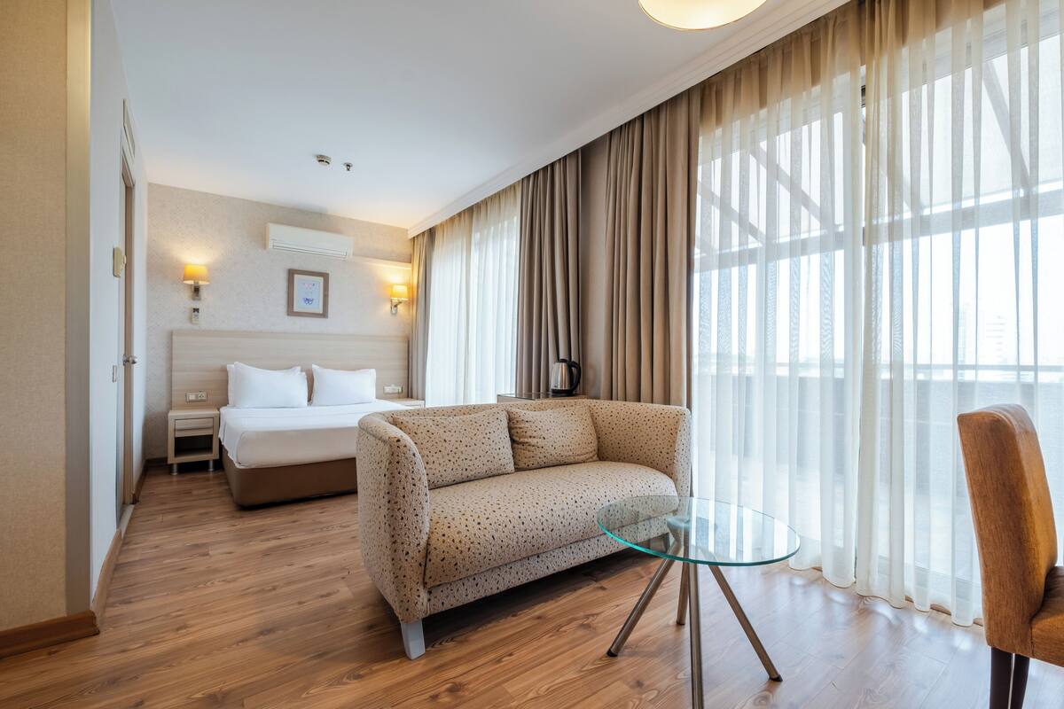 Una habitación de hotel con cama, sofá, una mesita de vidrio y una silla. Hay cortinas en las ventanas y una lámpara de pared.
