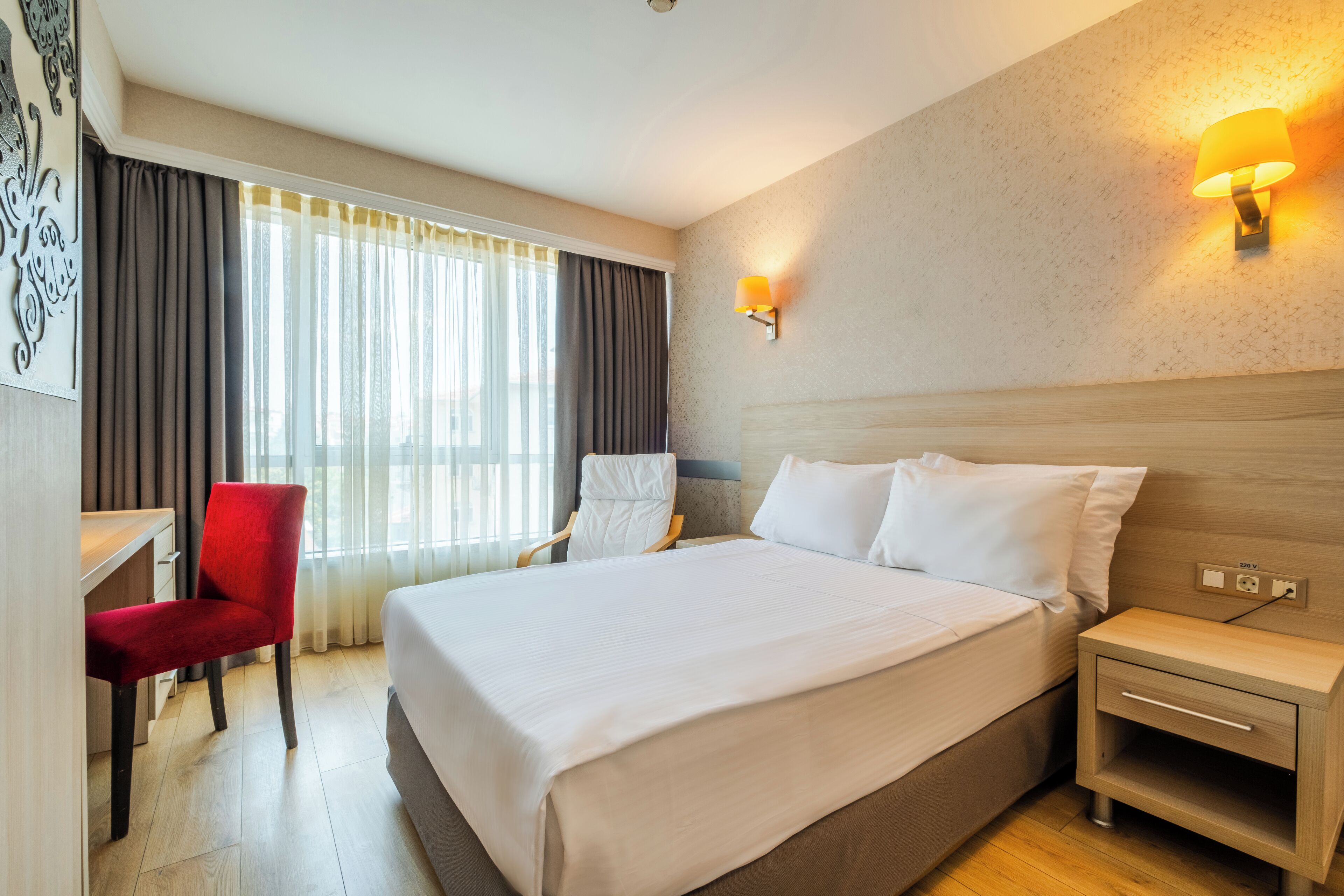 A hotel room with a large bed, a desk with a chair, a nightstand, and a wall-mounted lamp.