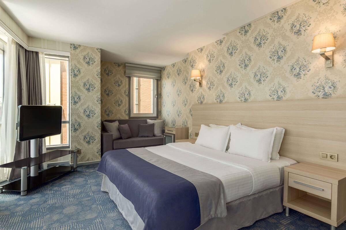 Habitación de hotel con una cama grande, un televisor sobre un soporte, un sofá y una mesita de noche con una lámpara.