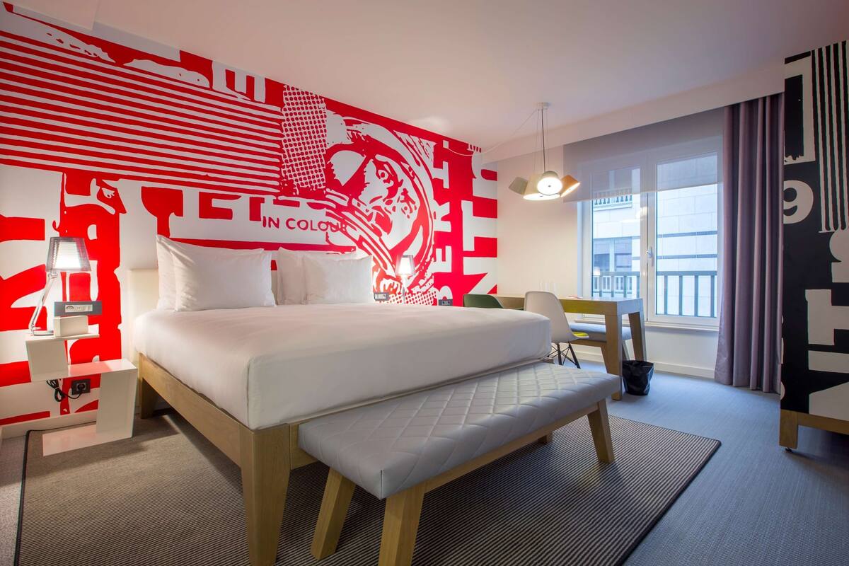 Quarto de hotel moderno com uma cama grande, uma mesa de trabalho e vista para a cidade.