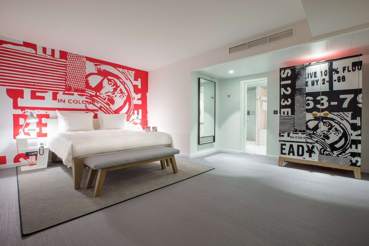 Quarto moderno com uma cama grande, um banco e uma parede com arte abstrata em vermelho e branco.