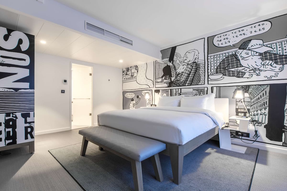 Quarto de hotel moderno com uma cama grande, um banco e um mural na parede.
