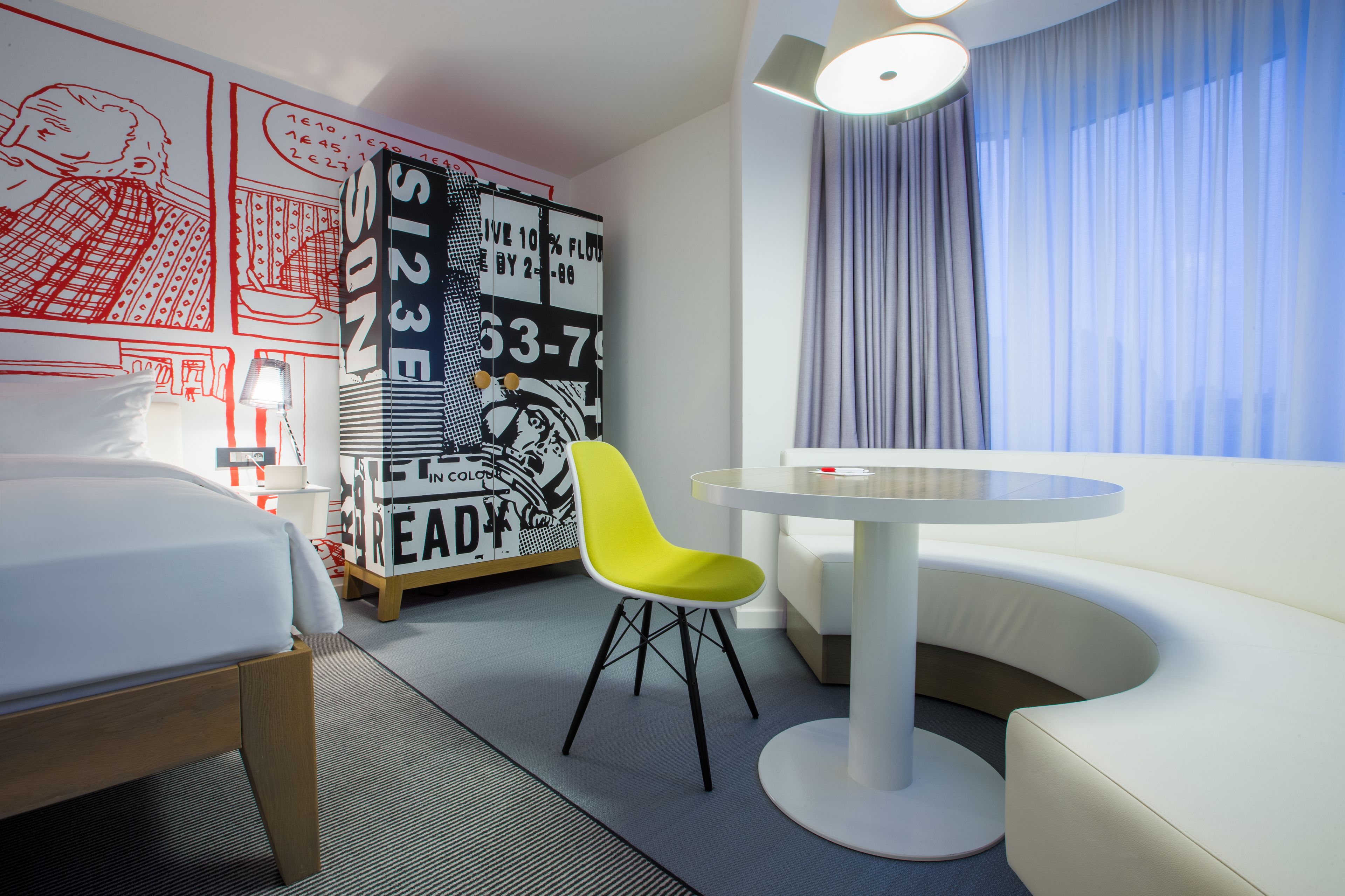 A hotel room with a bed, a bright yellow chair, a round white table, and a wall with a large poster.